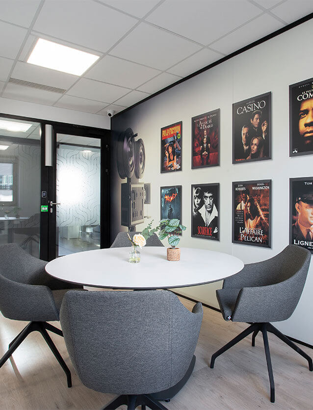 Photo du coin réunion dans le bureau de direction du siège de CEGELEM, avec une table ronde blanche, 4 chauffeuses grises et plusieurs affiches de cinéma