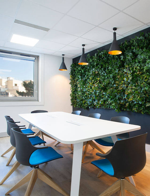 Photo de l'une des salles de réunion avec un mur entièrement végétalisé, une table de réunion blanche avec 2 boîtiers sur le plateau ainsi que 8 chaises en polypro noir, piètement pyramidal en bois et assise tapissée bleue